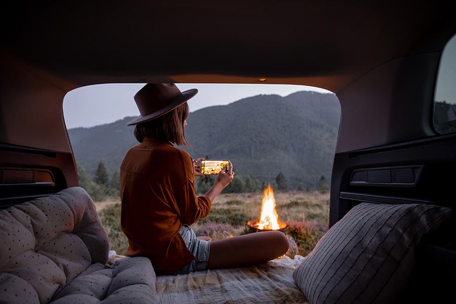 young woman camping in the back of her SUV, taking a picture of the campfire with her phone